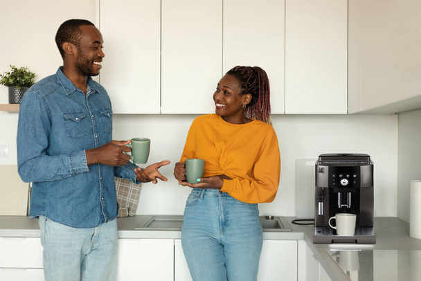 Young african american spouses drinking morning coffee and talking while standing near coffee machine in kitchen interior. Black couple enjoying time at home - Foto, immagini