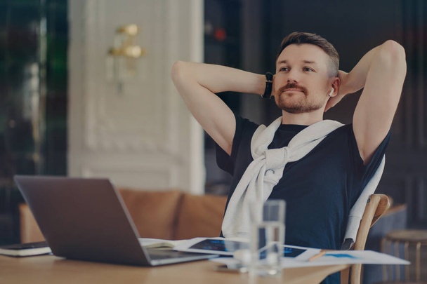 Pensive brodaty człowiek zastanawia się nad zadaniem trzyma ręce za głową siedzi na pulpicie z nowoczesnych gadżetów myśli o pracy na odległość koncentruje się na odległość nosi t koszulę sweter związany na ramionach - Zdjęcie, obraz