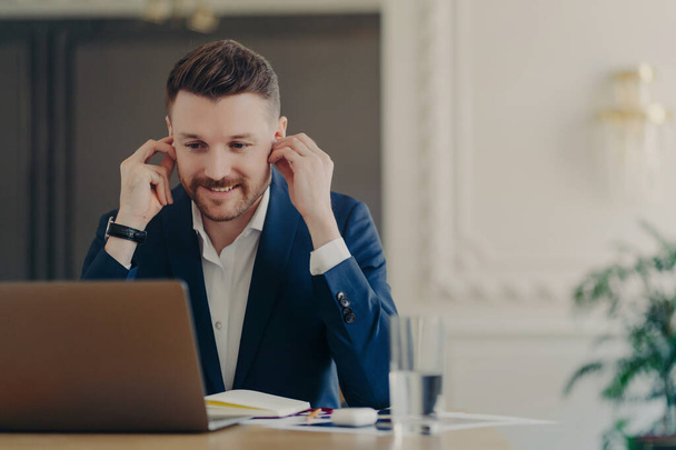 Gerente ejecutivo sonriente hombre de negocios que usa traje azul formal que tiene reunión en línea con los empleados que usan auriculares inalámbricos mientras está sentado frente a la computadora portátil en el escritorio de la oficina - Foto, Imagen