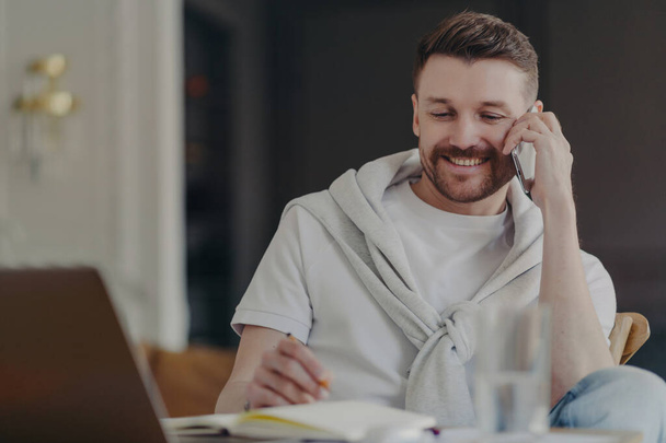 Attractive smiling male freelancer or businessman talking on phone while sitting at his work desk in front of laptop at home office making notes, wearing tshirt and sweater over his shoulders - Fotoğraf, Görsel
