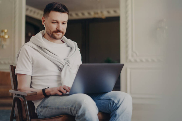 Young happy man freelancer in casual clothes wearing earphones using laptop while working from home, listening to music and looking at computer screen while sitting at modern home office interior - 写真・画像