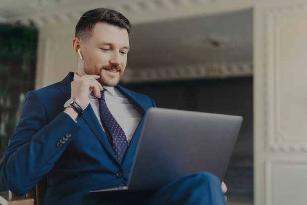 Confident handsome businessman in formal suit wearing wireless earphones looking at laptop screen during online business meeting, discussing project with colleagues, working in luxury hotel or office - Foto, imagen