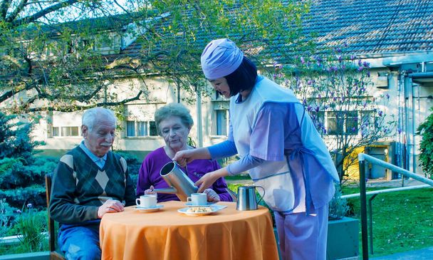 Fiatal ázsiai pincérnő, aki idős nyugdíjasoknak szolgál fel reggelit egy kórházi rehabilitációs kertben.. - Fotó, kép