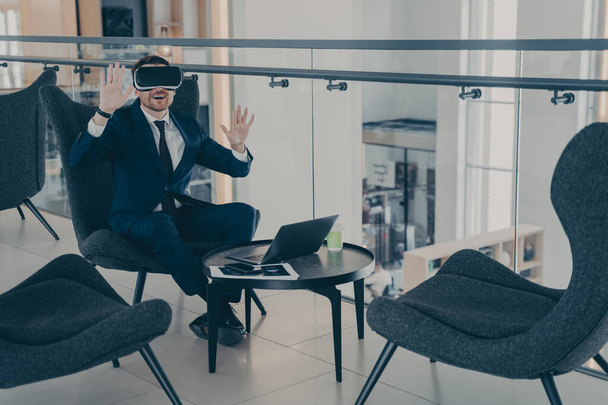 VRヘッドセットのビジネスマンは、コーヒーブレイク中にオフィスセンターのフロントエリアに座って、仮想現実のオブジェクトと対話します。ジェスチャーを使ってデジタルシミュレーションを行う会社のCEO - 写真・画像