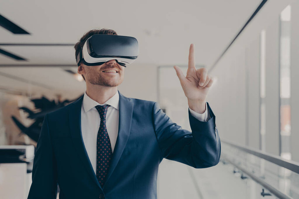 Cheerful businessman dressed formally using vr headset at work, wearing virtual reality glasses and working in augmented reality, pointing at virtual objects with finger. Futuristic technology in business - Foto, immagini