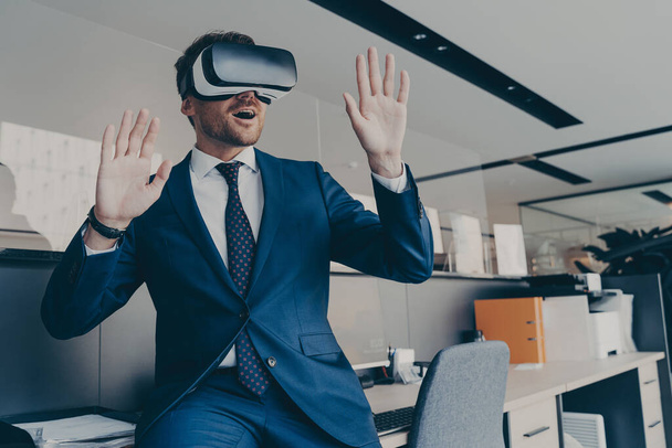 Úžasný muž ve VR headsetu interaguje s virtuálními objekty nebo rozhraním, sedí na okraji kancelářského stolu. Nadšený podnikatel dělá 3D obchodní prezentaci nebo setkání s brýlemi virtuální reality - Fotografie, Obrázek