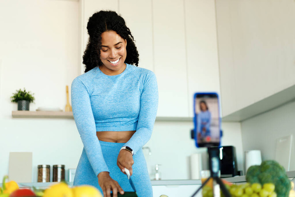 Ευτυχισμένη Αφρικανή Αμερικανίδα κυρία με αθλητικά ρούχα που ηχογραφεί βίντεο σε κάμερα smartphone, ετοιμάζοντας φρέσκια σαλάτα στην κουζίνα. Γυναίκα κοπής λαχανικών στο τραπέζι - Φωτογραφία, εικόνα