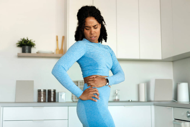 Dissatisfied fit black lady holding aching hip, has kidney inflammation, touching location of paim, wearing sport clothes, having sporty shaped body - Foto, afbeelding