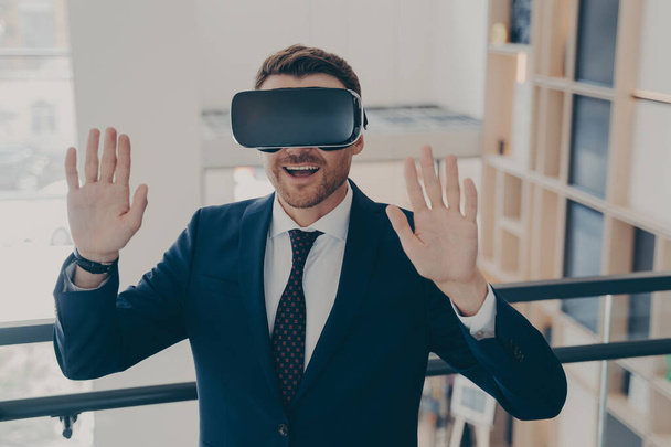 Lächelnder Büroangestellter im Anzug mit 3D-Brille genießt Cybertempo-Erfahrung mit erhobenen Armen, überraschter aufgeregter Geschäftsmann gestikuliert und berührt Objekt in virtueller Realität mit VR-Headset - Foto, Bild