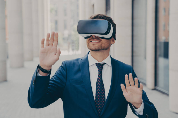 Young excited man in suit standing on city street and testing VR glasses or 3d goggles, impressed cheerful businessman using VR technology for the first time, gesturing and touching objects in air - Photo, Image