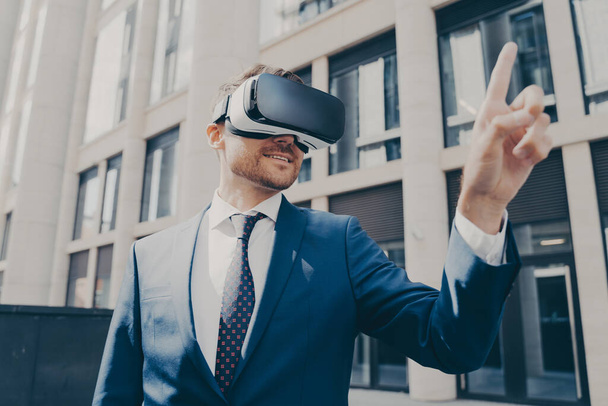 Young smiling handsome male enterpruer with stubble, dressed in formal attire, wearing portable VR goggles, trying to touch something in virtual reality with his finger, standing alone on city street - Foto, immagini
