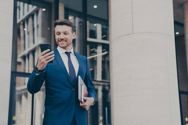 Young positive handsome businessman in elegant outfit using mobile phone, texting sms or checking email while standing outdoors against office building after hard working day, holding laptop in hand - Foto, Bild