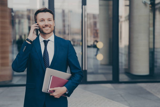 Happy male company worker leaving office building holding notebook and laptop while talking on phone with his colleague, sharing good news about recent events, blurred background - Photo, Image