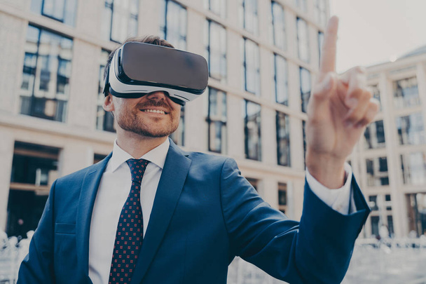 Hivatalos kék öltönyös üzletember VR szemüveggel odakint, kölcsönhatásban a virtuális világgal, megpróbál megérinteni valamit előtte, miközben mosolyog, elmosódott irodaházak a háttérben - Fotó, kép