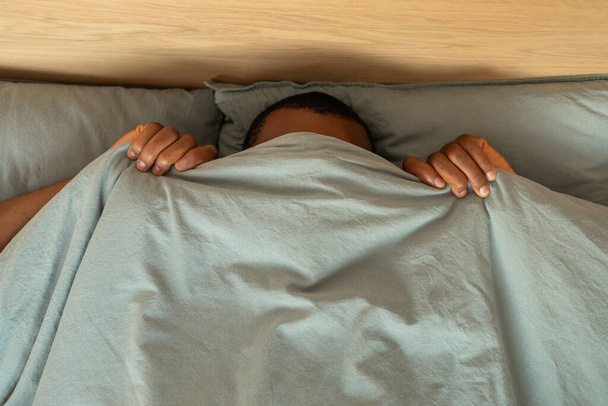 Top View Of Black Man Hiding Covering Face With Blanket Lying In Bed In Modern Bedroom At Home. Anxiety And Fear, Sleeping Routine Concept. High Angle Shot - Zdjęcie, obraz