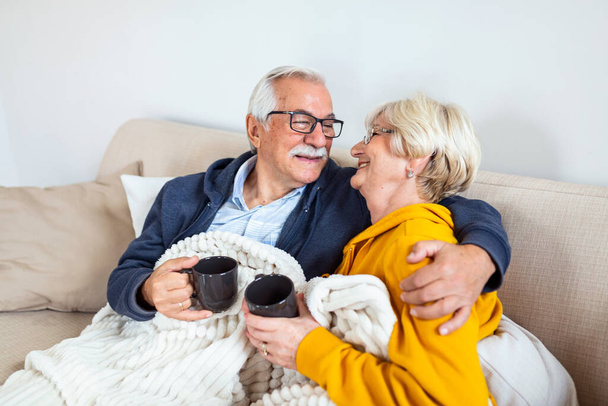 Happy old couple sitting on the sofa. Feeling cozy,drinking coffee or tea - Foto, afbeelding