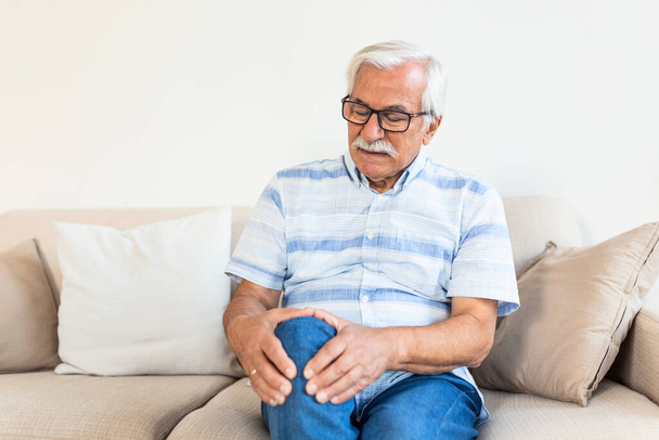 Пожилой мужчина сидит дома на диване и трогает колено от боли.. - Фото, изображение