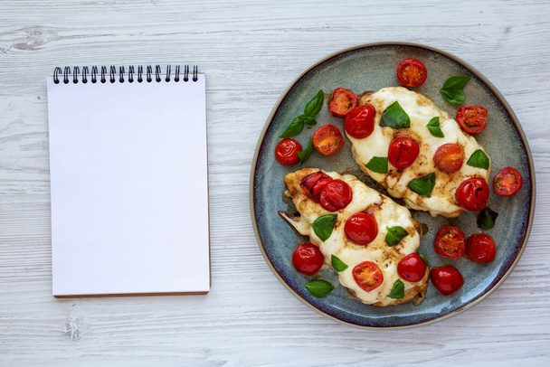 Homemade Caprese Chicken Parmesan with tomato, mozzarella and basil on a Plate, blank notepad, top view. Flat lay, overhead, from above. - Photo, image