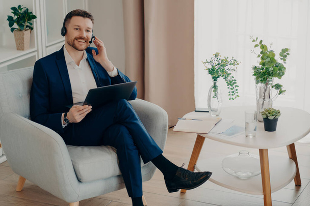 Концепция онлайн встреч. Улыбающийся элегантный мужчина в костюме, сидящий в кресле в гостиной и работающий удаленно на ноутбуке, бизнесмен регулирует гарнитуру во время видеочата - Фото, изображение
