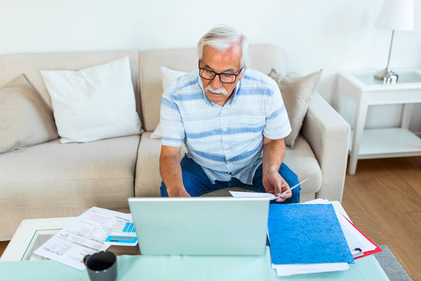 Ηλικιωμένος άνδρας που κάθεται στον καναπέ στο σπίτι σχεδιασμό του προϋπολογισμού ελέγχει τους λογαριασμούς των λογαριασμών, τον υπολογιστή, τα έγγραφα και αριθμομηχανή στο τραπέζι του καφέ, κλείστε περικοπεί εικόνα έννοια, γέρος διαχειρίζεται την έννοια των οικογενειακών δαπανών - Φωτογραφία, εικόνα