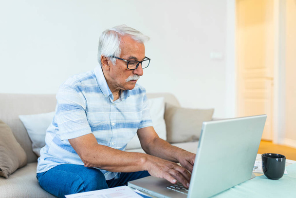 Starší podnikatel sedí na pohovce a pracuje na počítačovém bezdrátovém zařízení pomocí aplikace e-bankingu, platí účty online, e-mail řeší problémy distančně, starší generace a moderní technologie - Fotografie, Obrázek