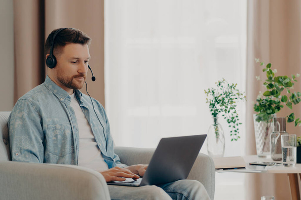 Young attentive bearded man in headset typing on laptop during online conference while sitting in armchair at home, male freelancer working remotely. Remote job concept - Valokuva, kuva