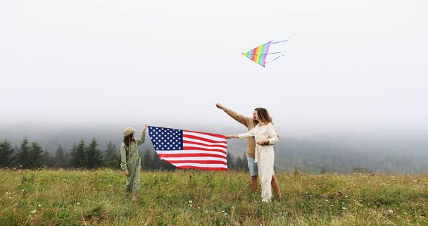 Patriotic holiday in United States of America. Happy family, parents and daughter child girl with American flag in nature on foggy mountains. The USA celebrate 4th of July - Photo, Image