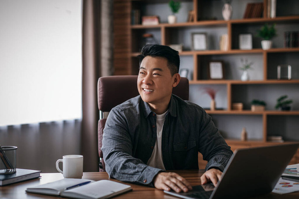 Happy mature chinese man at table working with laptop in home office interior and looking at empty space. Businessman analyzes sales for successful business, trade, development, good ad and offer - Zdjęcie, obraz