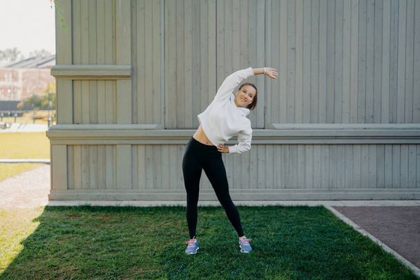 Sporty lifestyle wellness and weight loss concept. Active woman in sportwear trains outdoors leans left stands on green grass warms up before workout has active life enjoys regular workout in open air - Foto, Bild