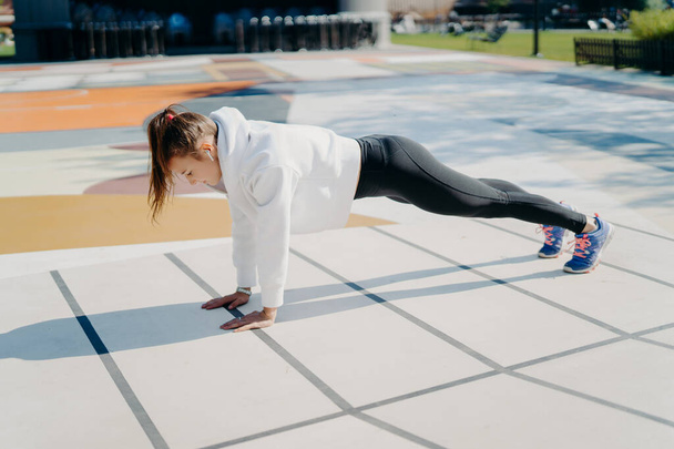Full length shot of active young woman with pony tail stands in plank focused down dressed in active wear poses outdoors being motivated gets stronger demonstrated endurance and determination - Foto, Bild