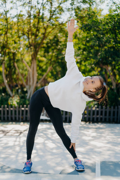 Vertical shot of glad sportswoman with combed pony tail leans aside keeps one arm raised up does exercises outdoors wears white hoodie leggings trainers relaxes on nature has active fitness training - Photo, Image