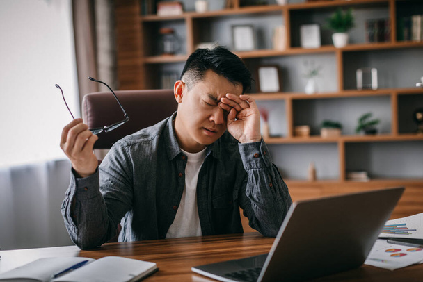 Sad tired adult chinese man takes off his glasses, rubs eyes and rests from work in home office interior. Overwork, problems in business, take break, stress and pressure, pain from incorrect posture - Photo, Image