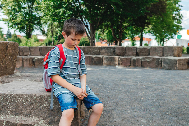 A sad schoolboy sits in the school yard with a backpack on his back. Childrens problems at school. The boy does not want to go to school. - Photo, image