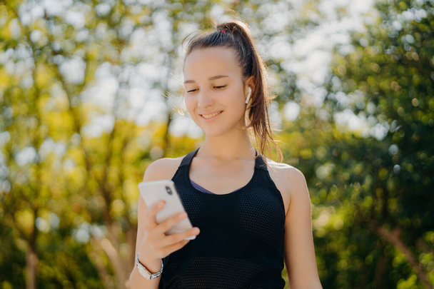 Відкритий постріл задоволеної спортивної жінки зосереджений на перевірці смартфона, скільки калорій вона спалила після вправ на бігу використовує спеціальний додаток, який слухає музику в навушниках має хвостову сорочку з поні
 - Фото, зображення