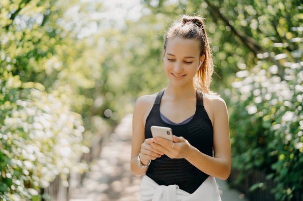 Sport lifestyle and workout concept. Young pleased sporty woman uses smartphone for chatting online and listening music rests after cardio training has combed pony tail dressed in active wear - Fotoğraf, Görsel