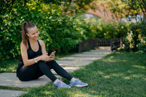 Pleased athletic woman in sportsclothes smiles cheerfully surfs mobile phone while listens music via wireless earphones takes break after sunny morning workout uses app for activity tracking - Photo, Image