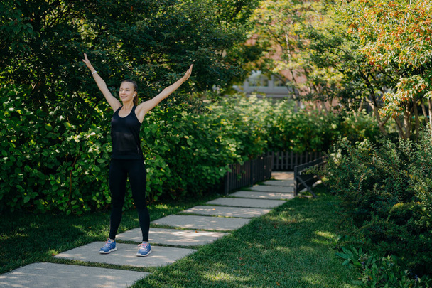 Full length shot of active sportswoman raises arms breathes deeply dressed in active wear has sport training in park during morning time listens music in earphones. Sporty lifestyle concept. - 写真・画像