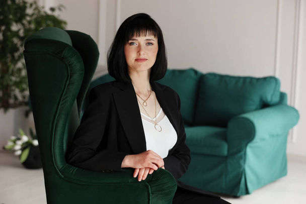 Beautiful business woman in black suit and white blouse is sitting on the green sofa at office, charming brunette female with short hair is looking in camera, girl is working indoors. - Foto, Imagem