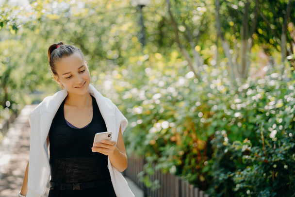 Pleased sporty woman with pony tail dressed in comfortable clothes focused at smartphone camera enjoys sport training outdoor walks in countryside checks message content. Lifestyle technology - Fotoğraf, Görsel