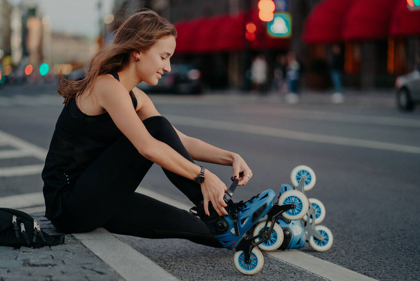 Outdoor shot of active woman laces rollerblades prepares for ride sits on road against busy city background dressed in black activewear enjoys rollerskating. Sporty lifestyle and recreation concept - Photo, image