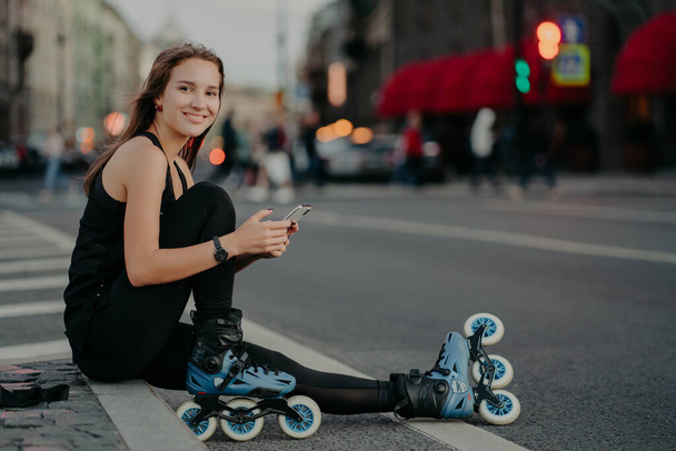 People outdoor activities and recreation concept. Horizontal shot of active slim woman being in good physical shape rides rollerblades uses smartphone sends text messages online poses outside - Fotó, kép