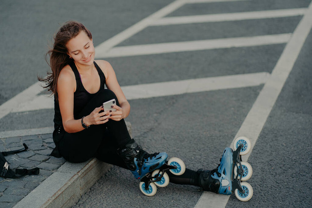 Top view of brunette active young woman uses smartphone for online communication poses on asphalt dressed in black active wear enjoys favorite hobby and free time. Extreme sport and rest concept - Photo, Image