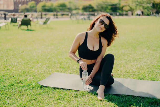 Outdoor shot of brunette slim young woman being in good physical shape trains on fitness mat drinks refreshing water wears sunglasses during sunny day cropped top and leggings. Workout concept - Photo, Image