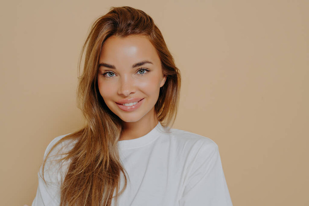 Portrait of beautiful female with long light brown straight hair gently smiling on camera wearing casual white tshirt while poses against pastel beige wall. Women beauty and positive emotions concept - Fotoğraf, Görsel