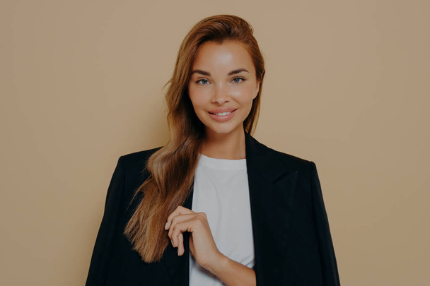 Friendly smiling brunette businesswoman with loose long hair, wears formal black jacket over white blouse, poses against beige background with blank space, happy to meet with business partners - Photo, Image
