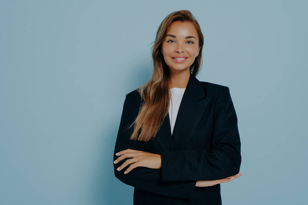 Studio shot of determined beautiful businesswoman in formal black suit standing proudly with crossed arms and smiling, posing against blue background with blank space. Positive body language - Photo, Image
