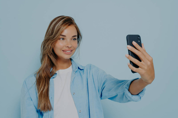 Selfie zamanı. Moda kıyafetleri içinde, uzun boyalı saçlarıyla modern cep telefonunda fotoğraf çeken açık mavi arka planda poz verirken büyüleyici ve cilveli bir genç kadın. - Fotoğraf, Görsel