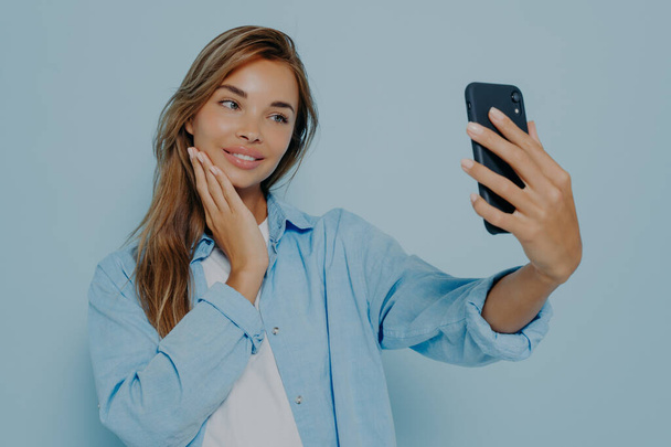 Łapię szczęśliwe chwile. Urocza młoda kobieta z selfie jej nowoczesny smartfon, delikatnie uśmiechnięta do aparatu podczas pozowania izolowane na jasnoniebieskim tle - Zdjęcie, obraz