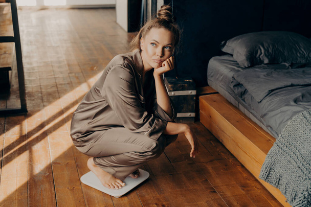 Egy fiatal, zaklatott nő képe, aki méri a súlyát, szatén pizsamába öltözve, elektronikus mérlegen guggolva kora reggel, miután felébredt a hálószobában. Fogyás és fogyókúra koncepció - Fotó, kép