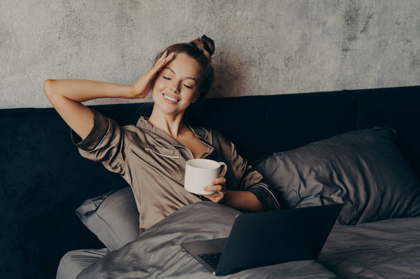 Okouzlující uvolněná mladá žena ležící doma v posteli s notebookem a s ranním šálkem kávy, sledování své oblíbené televizní show on-line na počítači, trávení volného času doma o víkendu - Fotografie, Obrázek